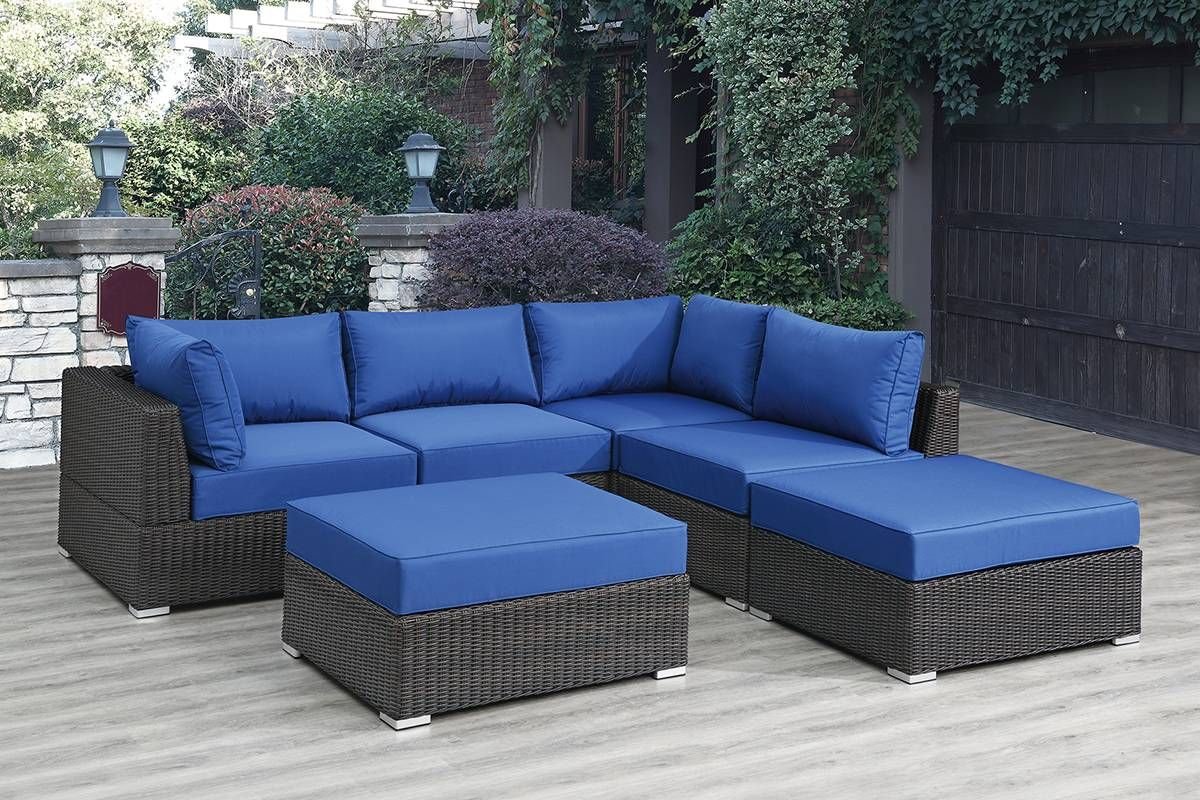 Outdoor patio set with blue cushions on dark wicker furniture, featuring a sectional sofa, two ottomans, and a low table, placed on a wooden deck amid greenery and a stone wall. 🔆 Ultimate 6-Piece Modular Outdoor Sectional Set – Transform Your Patio Today! 🌿